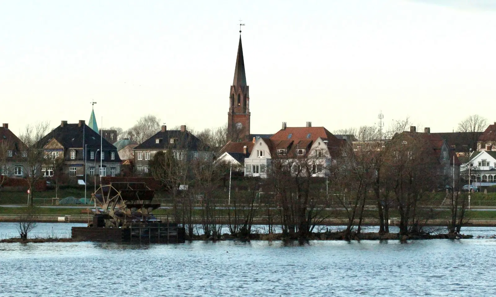 Fredrikstad sentrum sett fra Glomma, med tårnet på Fredrikstad domkirke, murvillaer og trær langs elvebredden i kveldslys